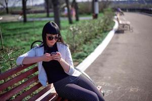 schattig vrouw met zonnebril en lang haar- is gebruik makend van mobiel apparaat genieten van zonnestralen en warm dag buitenshuis. portret van glimlachen brunette met smartphone in de park. foto