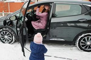 jong moeder met baby meisje opladen elektrisch auto in de werf van haar huis Bij winter. foto