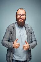 hij levensstijl van een geslaagd jong Mens met bril , baard, modieus denim jasje tonen duimen omhoog, heren emotioneel portret in studio foto