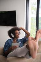 Afrikaanse Amerikaans vrouw Bij huis in stoel met tablet en hoofd telefoons foto