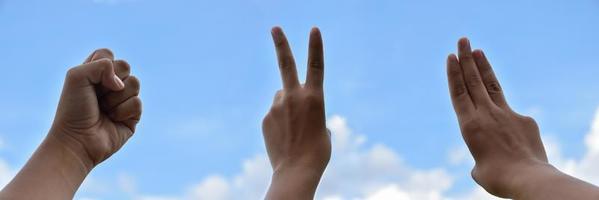 handen van menselijk in vuist, drie en twee vingers tonen Aan bewolkt en blauwe lucht achtergrond. zacht en selectief focus Aan vingers. foto