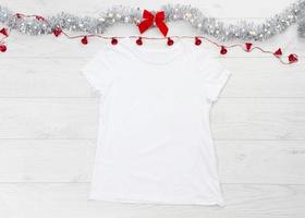 dichtbij omhoog wit blanco sjabloon t overhemd met kopiëren ruimte en Kerstmis vakantie concept. top visie mockup t-shirt en rood boog Aan wit houten achtergrond. gelukkig nieuw jaar decoraties accessoires. Kerstmis kleding foto