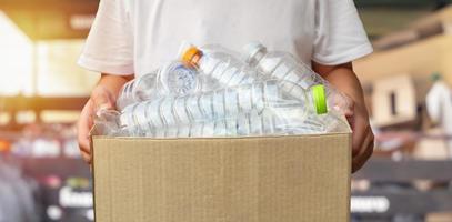 hand- Holding bruin recycle vuilnis doos met plastic flessen met klein recycle station achtergrond foto