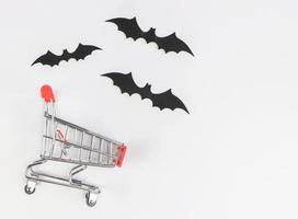 vlak leggen van boodschappen doen kar, halloween decoraties, zwart papier vleermuizen Aan wit achtergrond met kopiëren ruimte. halloween vakantie boodschappen doen concept. foto