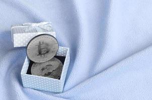 de zilver bitcoin leugens in een klein blauw geschenk doos met een klein boog Aan een deken gemaakt van zacht en pluizig licht blauw fleece kleding stof met een groot aantal van Verlichting vouwen foto