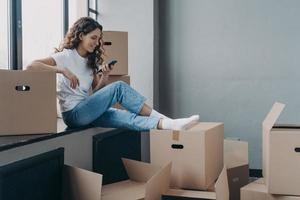 meisje Holding telefoon zittend Aan vensterbank met dozen selecteert in beweging onderhoud, planning huis vernieuwing foto