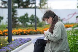 een meisje zit Aan een bank Aan de straat met documenten in haar handen en gebruik makend van haar telefoon foto
