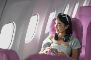 jong vrouw zittend met telefoon Aan de vliegtuig stoel in de buurt de venster gedurende de vlucht in de vliegtuig foto
