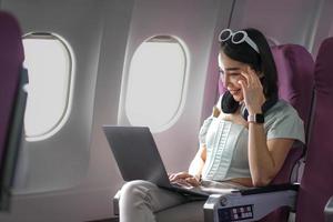 Aziatisch jong vrouw gebruik makend van laptop zittend in de buurt ramen Bij eerste klasse Aan vliegtuig gedurende vlucht, reizen en bedrijf concept foto