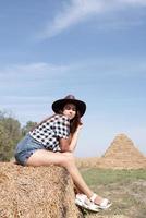 mooi vrouw in plaid overhemd en cowboy hoed resting Aan hooiberg tegen lucht achtergrond foto