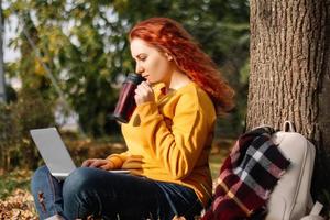 jong vrouw drankjes koffie en werken gebruik makend van laptop in natuur. afgelegen werk Aan de internetten. rust uit en ontspanning Aan herfst zonnig dag. foto