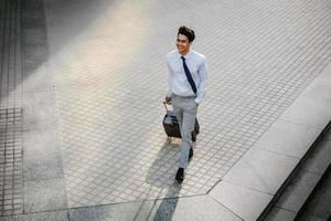 gelukkig jong zakenman wandelen met koffer in de stad. modern mannetje in bedrijf reis. hoog hoek visie. vol lengte foto