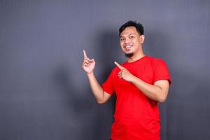 Aziatisch jong Mens in rood t-shirt richten met zijn vinger geïsoleerd Aan grijs achtergrond foto