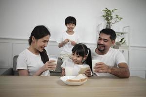 een gezond Aziatisch Thais familie, weinig kinderen, en jong ouders drinken vers wit melk in glas en brood vreugde samen Bij een dining tafel in ochtend, welzijn voeding huis ontbijt maaltijd levensstijl. foto