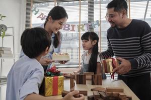 gelukkig Aziatisch Thais familie, jong kinderen verrast door verjaardag taart, geschenk, blazen een kaars, en vieren partij met ouders en broers en zussen samen Bij dining tafel, welzijn huiselijk huis speciaal evenement. foto