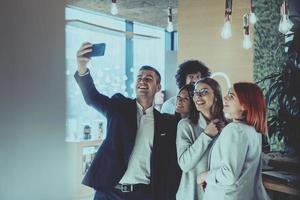 groep van vrolijk collega's nemen selfie en gebaren terwijl staand in de modern kantoor. foto