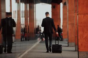 gaan naar luchthaven terminal. zelfverzekerd zakenman reiziger wandelen Aan stad straten en trekken zijn koffer drinken koffie en sprekend Aan smartphone foto