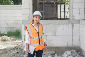 portret van een vrouw ingenieur Bij de gebouw plaats op zoek Bij de camera en glimlachen Holding een tablet vervelend een oranje veiligheid hesje en harde hoed foto