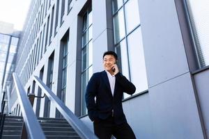 geslaagd Aziatisch zakenman pratend Aan de telefoon in de buurt de kantoor centrum glimlachen foto