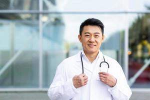 portret van een vrolijk Aziatisch dokter Mens glimlachen Aan de achtergrond van een modern kliniek buitenshuis foto