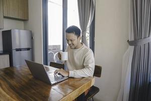 een jong Aziatisch Mens besteedt zijn tijd Bij huis zittend in de dining kamer gelukkig werken Aan zijn laptop terwijl Holding een mok foto