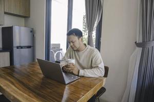 een jong Aziatisch Mens besteedt zijn tijd Bij huis zittend in de dining kamer werken Aan zijn laptop voelen gefrustreerd foto