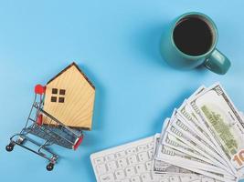 vlak lay-out van houten huis model- in boodschappen doen karretje, computer toetsenbord, bankbiljetten en blauw kop van zwart koffie Aan blauw achtergrond met kopiëren ruimte, huis aankoop concept. foto