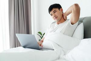 Aziatisch Mens in wit t-shirt houdende Aan bed gebruik makend van laptop computer hebben nek pijn. foto