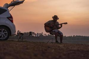 een Aziatisch Mens zit in de terug van zijn auto terwijl spelen een gitaar in een klein camping Bij zonsondergang. foto