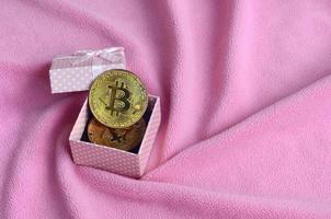 de gouden bitcoin leugens in een klein roze geschenk doos met een klein boog Aan een deken gemaakt van zacht en pluizig licht roze fleece kleding stof met een groot aantal van Verlichting vouwen foto