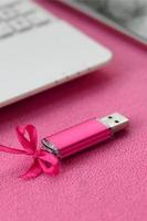 briljant roze USB flash geheugen kaart met een roze boog leugens Aan een deken van zacht en harig licht roze fleece kleding stof naast naar een wit laptop en smartphone. klassiek vrouw geschenk ontwerp voor een geheugen kaart foto