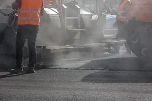 mannen werken met asfalteren bestratingsafwerkmachine machine gedurende weg straat repareren werken Bij dag licht met rook en stoom- in de lucht foto
