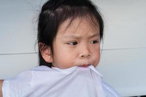 portret schattig Aziatisch meisje trekt haar mond over- halsband van haar t shirt. uitdrukken turbulent of in verlegenheid gebracht emoties. kind fronste, maken verdacht of ongemakkelijk uitdrukking. kind 5 jaren oud. foto