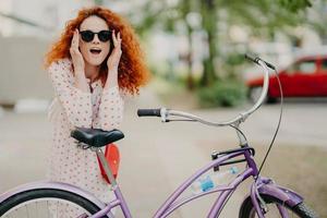 gelukkige vrouw heeft krullend foxy haar, leunt op het zadel van haar fiets, heeft een buitenpromenade tijdens zonnige zomerdag, draagt stijlvolle tinten en kleding, poseert over stedelijke omgeving. gezonde levensstijl en rust foto