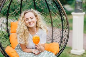 schattige krullende jonge vrouw met krullend borstelig haar, houdt een glas sinaasappelcocktail vast, poseert in een hangende buitenstoel, drukt positiviteit uit, heeft een prettig gesprek met de gesprekspartner. vrije tijd concept foto