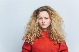 foto van ontevreden ongelukkige jonge vrouw met krullend borstelig blond haar, fronst gezicht in verbijstering, draagt casual kleding, modellen tegen witte studio achtergrond. mensen en teleurstelling concept