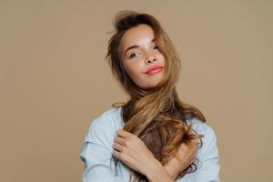 portret van doordachte mooie vrouw kijkt opzij, denkt aan iets aangenaams, steekt haar over nek, draagt make-up, stijlvol shirt, geïsoleerd op bruine achtergrond. mensen, schoonheid, levensstijl foto