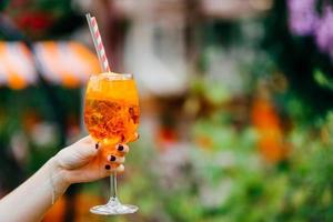 schot van de hand van de vrouw met manicure houdt een vers zomerdrankje in een wijnglas. oranje koude cocktail met ijs om te drinken tegen onscherpe achtergrond foto
