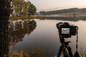 zwart digitaal camera Aan statief het schieten vroeg mistig ochtend- landschap Bij herfst meer met selectief focus foto