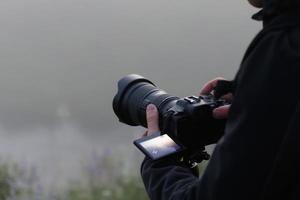 onherkenbaar fotograaf het schieten nevelig buitenshuis tafereel met hedendaags zwart digitaal camera Aan een statief met omdraaien scherm foto