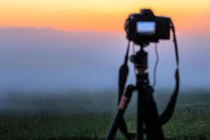 zwart digitaal camera Aan statief het schieten mistig ochtend- landschap Bij zomer met selectief focus foto