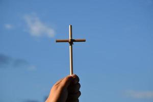 klein houten kruis Holding in hand- met bewolkt en blauw lucht achtergrond, concept voor liefde, hoop, waarheid, vertrouwen, van mening zijn in Jezus, zacht en selectief focus. foto