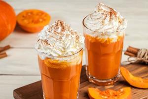 pittig latte met pompoen en geslagen room Aan houten achtergrond. heet koffie in glas mok en herfst bladeren. foto