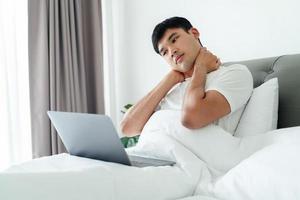 Aziatisch Mens in wit t-shirt houdende Aan bed gebruik makend van laptop computer hebben nek pijn. foto