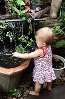 weinig meisje met blond haar- in zonnejurk is staand in tuin met pot planten en waterval en spelen met Afdeling van boom. foto