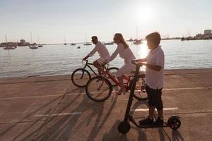 gelukkig familie genieten van een mooi ochtend- door de zee samen, ouders rijden een fiets en hun zoon rijden een elektrisch scooter. selectief focus foto