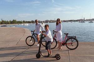 gelukkig familie genieten van een mooi ochtend- door de zee samen, ouders rijden een fiets en hun zoon rijden een elektrisch scooter. selectief focus foto