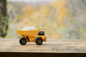 een klein geel speelgoed- vrachtauto is geladen met een steen van wit zout. een auto Aan een houten oppervlakte tegen een achtergrond van herfst Woud. extractie en vervoer van zout foto