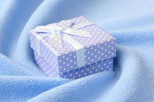 een klein geschenk doos in blauw met een klein boog leugens Aan een deken van zacht en harig licht blauw fleece kleding stof met een veel van Verlichting vouwen. inpakken voor een geschenk naar uw lief vriendin foto