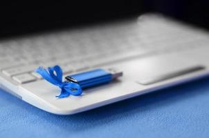 briljant blauw USB flash geheugen kaart met een blauw boog leugens Aan een deken van zacht en harig licht blauw fleece kleding stof naast naar een wit laptop. klassiek vrouw geschenk ontwerp voor een geheugen kaart foto